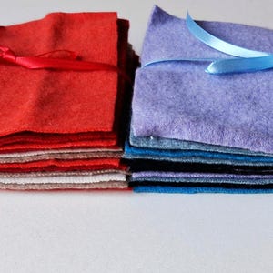 May include: Two stacks of felt squares, one red and one purple, are tied with ribbons. The red stack has a red ribbon, and the purple stack has a blue ribbon. The felt squares are in various colours, suitable for crafting.