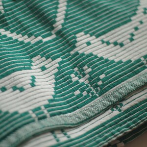 Op de afbeelding: Een close-up van een geweven stof in groen en wit met een geometrisch patroon. De stof is over zichzelf gevouwen, waardoor een textuur ontstaat.