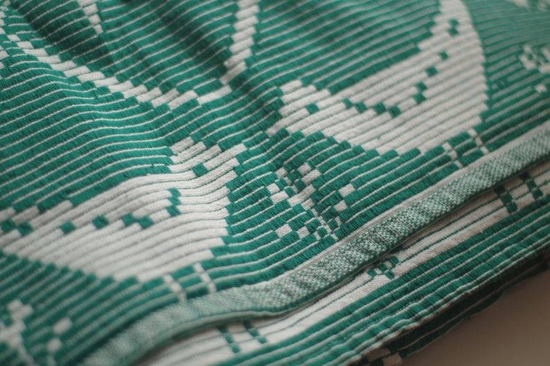 Op de afbeelding: Een close-up van een geweven stof in groen en wit met een geometrisch patroon. De stof is over zichzelf gevouwen, waardoor een textuur ontstaat.
