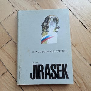May include: A book titled "Stare Podania Czeskie" by Aloizy Jirasek. The cover features a stylized illustration of a face with red, white, and blue accents. The book is light gray and rests on a wooden surface.