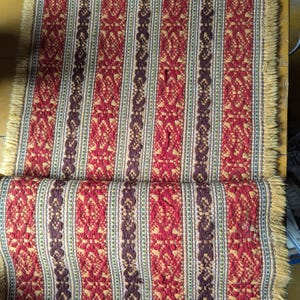 Puede incluir: Textil doblado con un patrón vertical repetido de motivos florales rojos, marrones y amarillos. Los bordes están acabados con una franja en tonos rojos y amarillos. El textil es probablemente una alfombra o tela decorativa.