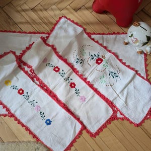 Puede incluir: Tres caminos de mesa de crochet blanco con ribete de crochet rojo y diseños florales bordados. Los caminos de mesa están apilados con el camino de mesa más grande en la parte inferior y el camino de mesa más pequeño en la parte superior.