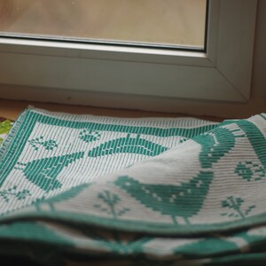 Op de afbeelding: Een witte en groene geweven keukendoek met een vogelontwerp. De handdoek is gevouwen en over een vensterbank gelegd.