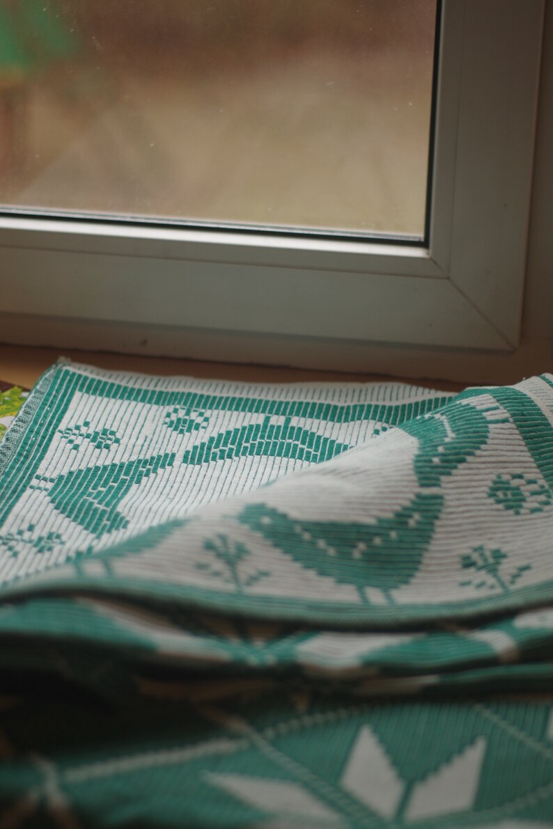 Op de afbeelding: Een witte en groene geweven keukendoek met een vogelontwerp. De handdoek is gevouwen en over een vensterbank gelegd.