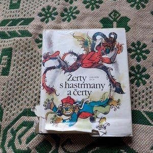 Peut inclure: Un livre vintage avec une couverture blanche ornée d'illustrations d'un diable rouge et d'une figure de clown. Le titre "Zerty s hastrmany a čerty" est imprimé en noir. La couverture du livre est légèrement usée.