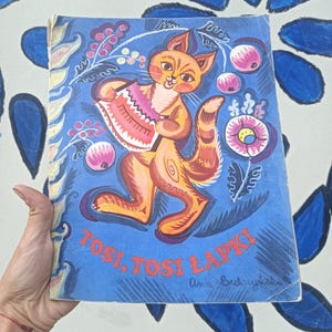 Puede incluir: Libro infantil vintage con una cubierta azul que presenta un gato de dibujos animados tocando el acordeón. El gato es naranja y rosa, rodeado de ilustraciones florales y frutales. El título "Tosi, Tosi Łapki" se muestra en rojo.
