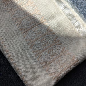 Puede incluir: Textil tejido de color crema con un patrón geométrico en un tono beige claro. La tela tiene un borde con flecos y una apariencia texturizada. El patrón es una serie de líneas verticales y horizontales, creando un diseño repetitivo.