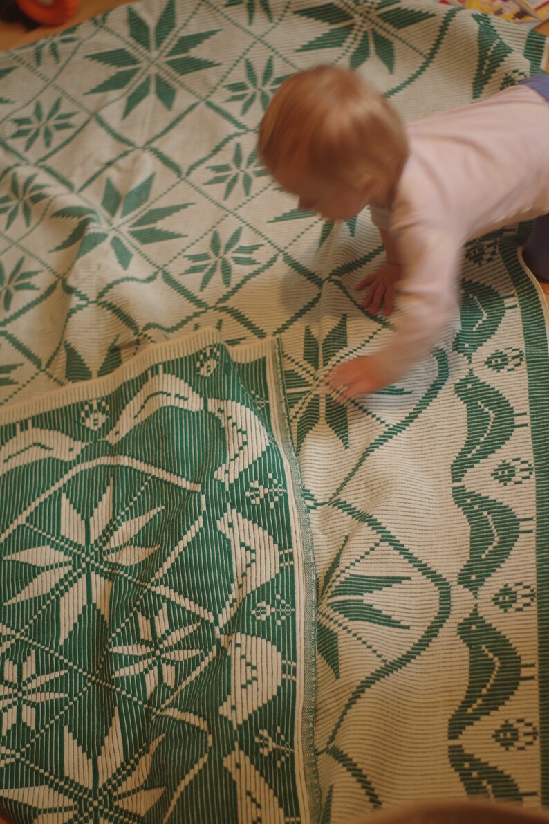 Op de afbeelding: Een geweven deken in groen en wit met een geometrisch patroon. De deken is dubbelgevouwen en een kind speelt erop.