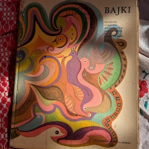 Può includere: Un libro vintage intitolato BAJKI con una colorata illustrazione di copertina raffigurante uccelli stilizzati nei toni dell'arancione, rosa, viola e verde. Il testo include TELEFON, LIMPOPO, MYJDODZIUR, SKRADZIONE, SLONCE, ZAMIESZANIE e WYDAWNICTWO RADUGA MOSKWA.