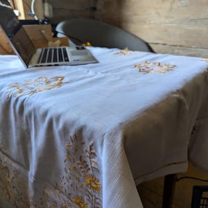 Puede incluir: Un mantel blanco con diseños florales amarillos bordados. El mantel está sobre una mesa, con un ordenador portátil visible en el fondo. El bordado presenta patrones detallados de flores y hojas.