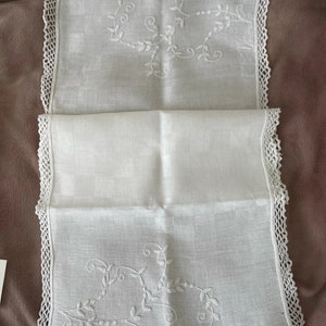Puede incluir: Un camino de mesa de lino blanco con bordes de ganchillo y diseños florales bordados. El camino de mesa está doblado por la mitad, mostrando el diseño en ambos lados.