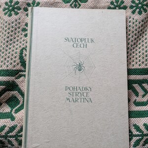 Peut inclure: Un livre vert clair avec le titre "SVATOPLUK CECH" et "POHADKY STRYCE MARTINA" imprimé sur la couverture. Un motif de toile d'araignée avec une araignée est au centre. Le livre est sur une surface verte et beige à motifs.