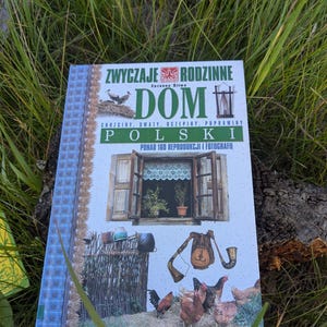 Può includere: Un libro intitolato "ZWYCZAJE RODZINNE DOM POLSKI" con illustrazioni di una finestra, galline e un corno. Il libro è appoggiato sull'erba, con un bordo a motivi blu e bianchi.