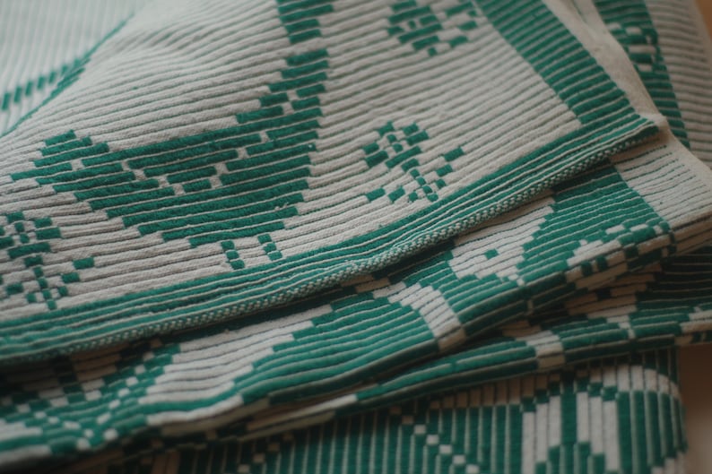 Op de afbeelding: Een close-up van een wit en groen geweven deken met een textuurpatroon. Het deken is gevouwen in meerdere lagen, waardoor het ingewikkelde ontwerp zichtbaar wordt.