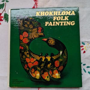 May include: A green book with the title "Khokhloma Folk Painting" in gold lettering. The cover features a black and red bird with floral and berry designs.