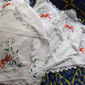 Puede incluir: Juego de cuatro servilletas de lino blanco con bordes de ganchillo y diseños florales bordados en rojo y verde.