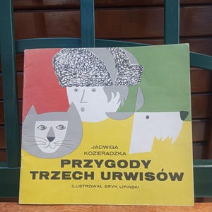 Puede incluir: Un libro infantil polaco vintage, titulado "Przygody Trzech Urwisów" (Aventuras de tres granujas). La portada presenta un gato, un perro y una persona con sombrero. El texto es de color amarillo.