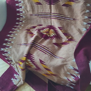 Può includere: Una coperta tessuta con un motivo geometrico in tonalità bordeaux, beige e oro. La coperta è piegata e drappeggiata su un divano bianco.