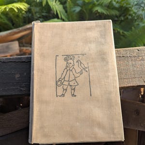Puede incluir: Un libro antiguo con una cubierta beige. La cubierta tiene una ilustración negra de una persona joven con un pájaro. La ilustración está dentro de un marco rectangular. El libro está sobre una superficie de madera, con follaje verde en el fondo.