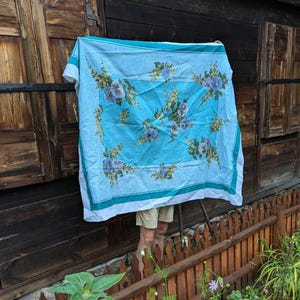 Puede incluir: Un mantel azul con un borde blanco y un patrón floral de flores azules y amarillas. El mantel está colgado en una pared de madera.