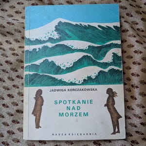 Puede incluir: Un libro vintage con una cubierta azul y verde que presenta olas estilizadas y el título "Spotkanie Nad Morzem". La cubierta también incluye siluetas de dos figuras y el nombre del autor, Jadwiga Korczakowska.