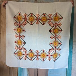 Può includere: Tovaglia di lino bianco con un bordo ricamato geometrico in rosso, arancione, giallo e nero.