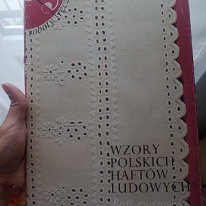 May include: A book cover with the text "Wzory Polskich Haftów Ludowych" and "roboty ręczne". The cover features a cream-colored fabric with white floral and geometric embroidery designs. The book has a red spine.