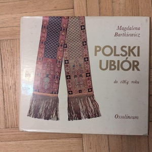 May include: A book cover featuring a detailed illustration of a traditional Polish textile design in shades of red, blue, and gold. The title "POLSKI UBIÓR" is prominently displayed, with the author's name and publisher's logo also visible.