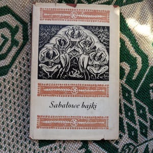 May include: Vintage book titled "Sabatowe bajki" with a black and white illustration of figures dancing in a forest. The cover has a cream background with orange decorative borders and text.