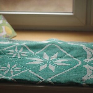 Op de afbeelding: Een turquoise en wit gepatroneerde deken met een ruitpatroon en sneeuwvlokkenontwerp. De deken is gevouwen en ligt op een houten oppervlak.