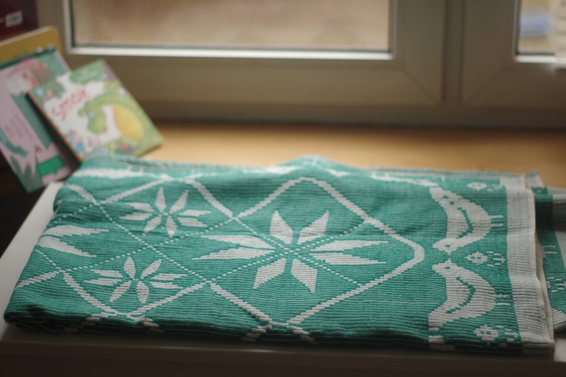 Op de afbeelding: Een turquoise en wit gepatroneerde deken met een ruitpatroon en sneeuwvlokkenontwerp. De deken is gevouwen en ligt op een houten oppervlak.