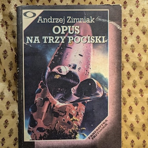 Peut inclure: Un livre de poche vintage intitulé "Opus Na Trzy Pociski" d'Andrzej Zimniak. La couverture présente une illustration de vaisseau spatial dans des tons de rouge, de marron et de noir. Le livre est posé sur un fond doré à motifs.