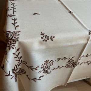 Puede incluir: Un mantel de color crema con diseños florales bordados en marrón. El mantel tiene un delicado patrón floral con hojas y flores.