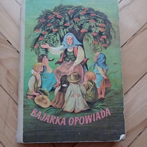 May include: A vintage children's book cover with a colorful illustration of a woman in a blue headscarf telling stories to a group of children under a tree with red berries. The book title is "BAJĄRKA OPOWIADA".
