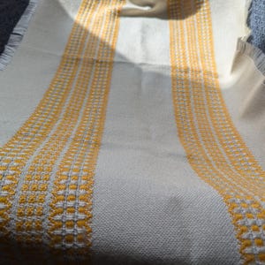 Puede incluir: Textil tejido de color crema con dos bandas verticales de patrones geométricos amarillos. El textil tiene un borde con flecos, probablemente una bufanda o una manta. La combinación de colores amarillo y crema crea un ambiente cálido.