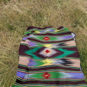 May include: A colorful woven blanket with a geometric pattern. The blanket features horizontal stripes in shades of brown, green, purple, and yellow. A diamond-shaped design is centered on the blanket, with red accents. The blanket is spread out on a grassy surface.