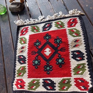 Puede incluir: Alfombra cuadrada tejida a mano con un campo central rojo con un patrón de diamantes negro con detalles en blanco y verde. El borde tiene patrones repetidos en rojo, verde, negro y beige. La alfombra tiene un borde negro y flecos blancos.