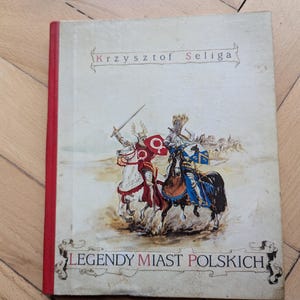 Puede incluir: Un libro con lomo rojo y portada descolorida que muestra ilustraciones de caballeros a caballo. El título es "Legendy Miast Polskich" y el autor es "Krzysztof Seliga". La portada presenta una ilustración histórica.