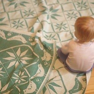 Op de afbeelding: Een jong kind zit op een wit en groen gestreept deken. Het deken heeft een geometrisch ontwerp met een herhalend patroon van bladeren en bloemen.