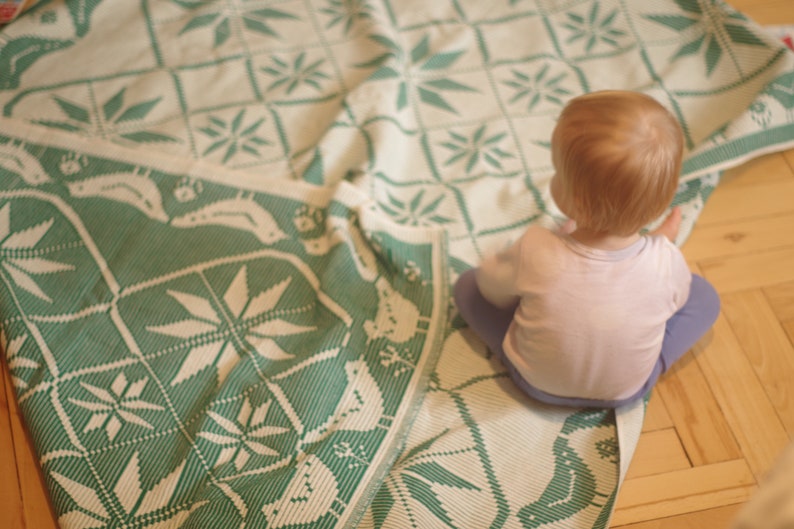 Op de afbeelding: Een jong kind zit op een wit en groen gestreept deken. Het deken heeft een geometrisch ontwerp met een herhalend patroon van bladeren en bloemen.