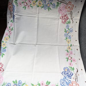 Puede incluir: Mantel blanco con diseños florales bordados en rosa, azul, amarillo y rojo. El bordado forma un borde a lo largo de los bordes, con patrones florales detallados y hojas verdes. La tela está doblada.