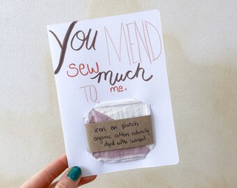 May include: A white card with the text "You mend sew much to me." written in brown and pink ink. A small pink and white fabric patch is attached to the card with the text "iron on patch organic cotton naturally dyed with cochineal" written on a brown label.