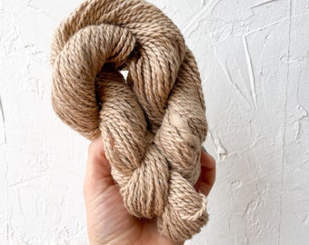 May include: A skein of natural brown yarn, made from a blend of cotton and linen, is held in a hand. The yarn is soft and textured, and would be perfect for knitting or crocheting.