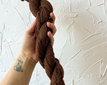 May include: A skein of brown yarn, held in a hand, against a white textured wall.