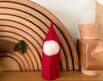 May include: A red felt gnome with a white felt face. The gnome is standing in front of a wooden rainbow toy.