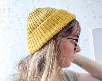 May include: A person wearing a mustard yellow beanie with a ribbed texture. The beanie is pulled down over their forehead and covers their ears. The person has long, blonde hair and is wearing glasses.
