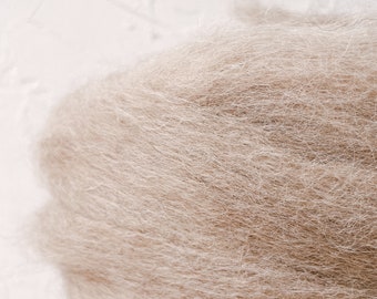 May include: A close-up of a pile of light brown wool fibers. The fibers are soft and fluffy, and they are arranged in a random pattern.
