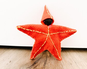 May include: A red felt star-shaped doll with a black felt head and a pointed red felt hat. The doll has yellow stitching around the edges.