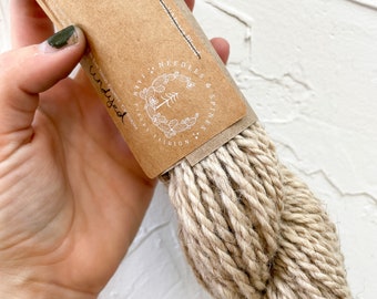 May include: A skein of undyed Navajo Churro wool yarn in a natural beige color. The yarn is packaged in a brown paper label with the text "100% Navajo Churro Wool 50g or 100g" and a logo that says "Needles & Spines Fashion".