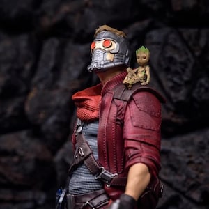 Könnte beinhalten: Eine rote und schwarze Actionfigur von Star-Lord aus Guardians of the Galaxy, die eine Maske mit rot leuchtenden Augen trägt. Eine kleine grüne Actionfigur von Groot sitzt auf seiner Schulter.